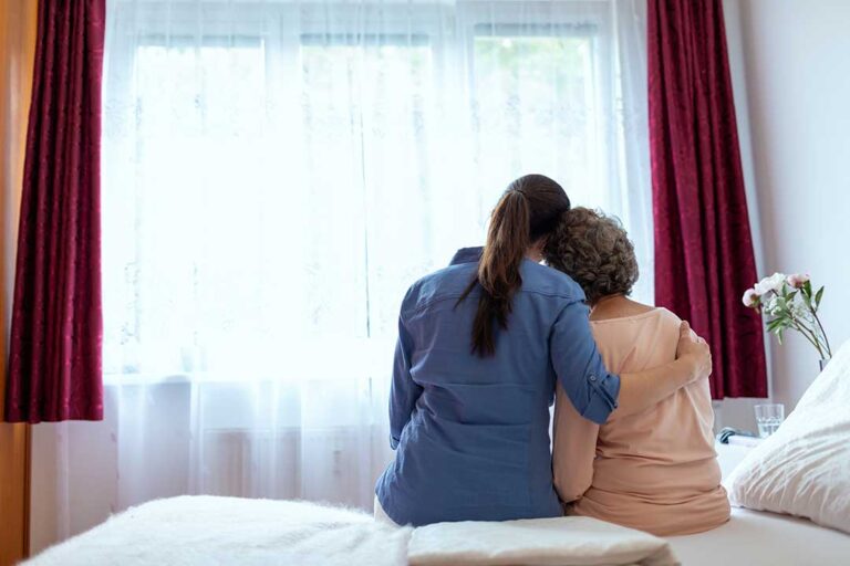 A social worker sits beside an older adult, listening and taking notes in a comfortable common area.