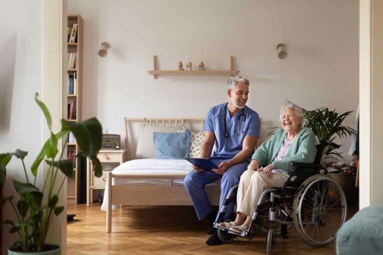 A caregiver assists an older adult with daily activities in a bright, home-like living space.